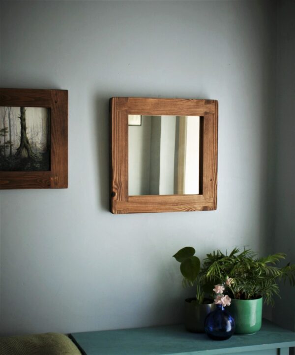 Square wooden frame mirror seen with plants and a picture frame.