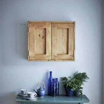 Wide kitchen cabinet in natural, rustic wood with 2 doors and blue crockery, glassware and leafy house plants on the table beneath.
