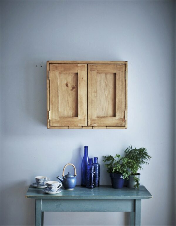 Wide kitchen cabinet in natural, rustic wood with 2 doors and blue crockery, glassware and leafy house plants on the table beneath.